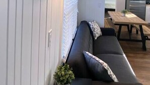 Een moderne woonkamer met een donkere bank, een houten wandplank met paddles, een tafeltje met een plant en een zicht op de keuken op de achtergrond.