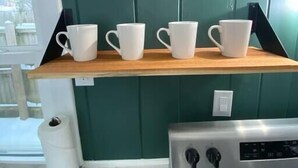 A kitchen with a green wall, a wooden shelf with four white mugs, a potted plant, and a stainless steel oven.