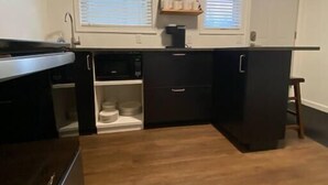 Een compacte keuken met donkere keukenkastjes, een inbouwoven en een houten aanrecht. Twee ramen met jaloezieën, een klein plankje boven de gootsteen en een deur aan de rechterkant.