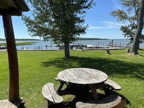 Una mesa de picnic con bancos, un área cubierta de césped y vista a un lago con muelles y barcos.
