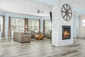 A spacious living room with a fireplace, large windows, and a ceiling fan.