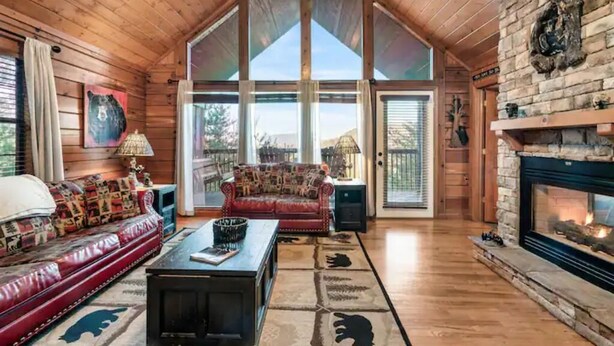 A log cabin interior with a large fireplace, a red sofa, a coffee table, and a view through the large windows. - King Bed, Pool Table, Hot tub, Vinyl Record Player, Covered Back Porch w VIEWS!! (Sevierville)