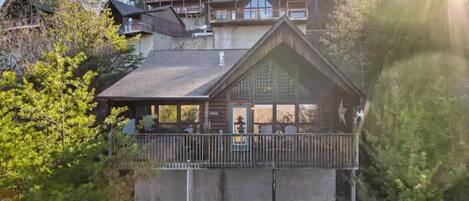 A wooden cabin with a balcony, surrounded by trees and other cabins in the background.