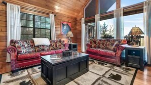 A spacious living room with wooden walls and large windows, featuring two red leather sofas with patterned cushions, a central coffee table, and a decorative lamp. - King Bed, Pool Table, Hot tub, Vinyl Record Player, Covered Back Porch w VIEWS!! (Sevierville)