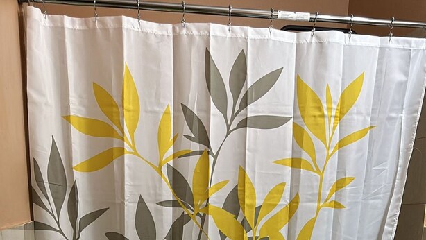 Een badkamer met een toilet, een gele mat, een zwarte prullenbak en een douchegordijn met bladmotief.