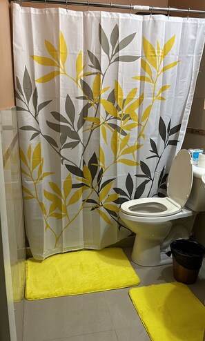 Un baño con inodoro, un tapete amarillo, un cesto de basura negro y una cortina de ducha con estampado de hojas.