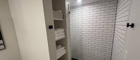 A bathroom with a white tiled shower, a toilet, and a towel rack with folded towels.