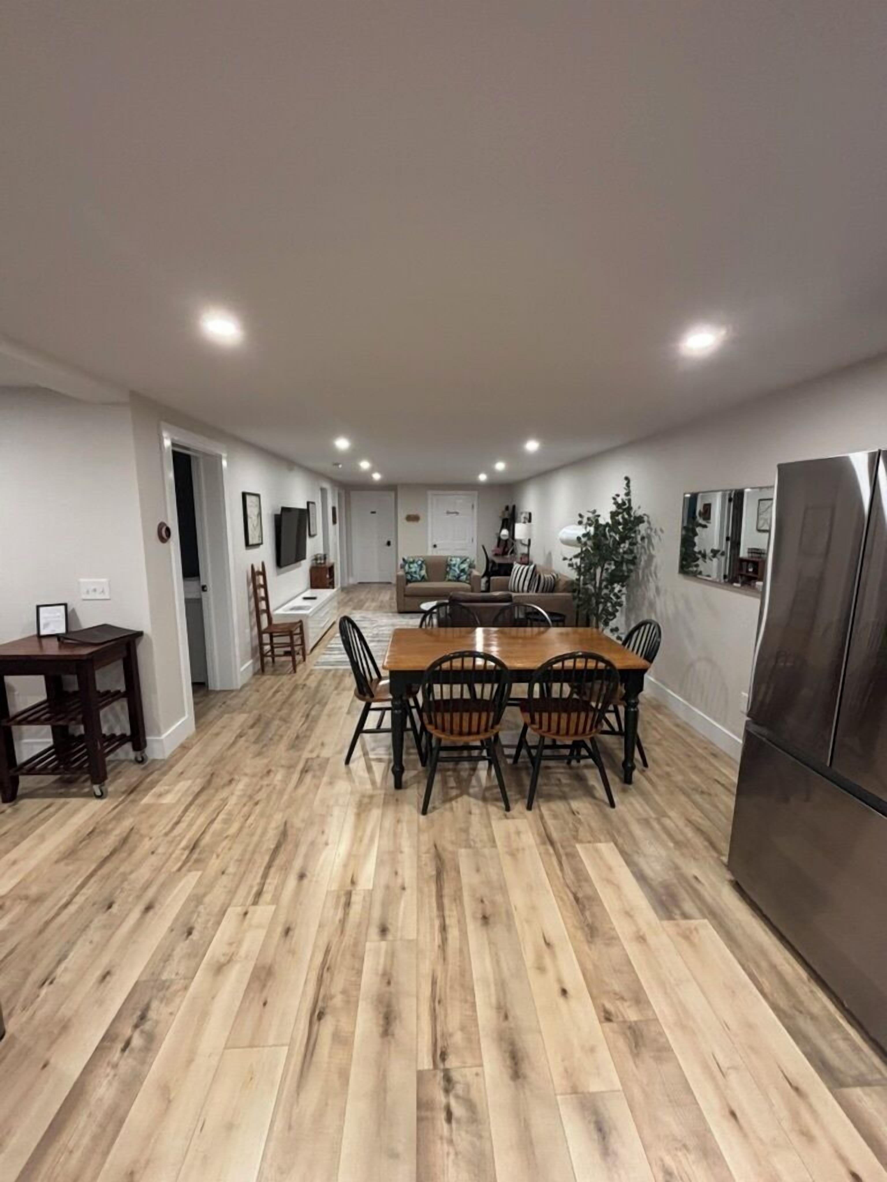 A spacious dining area with a wooden table and chairs, a refrigerator, and a kitchen in the background.
