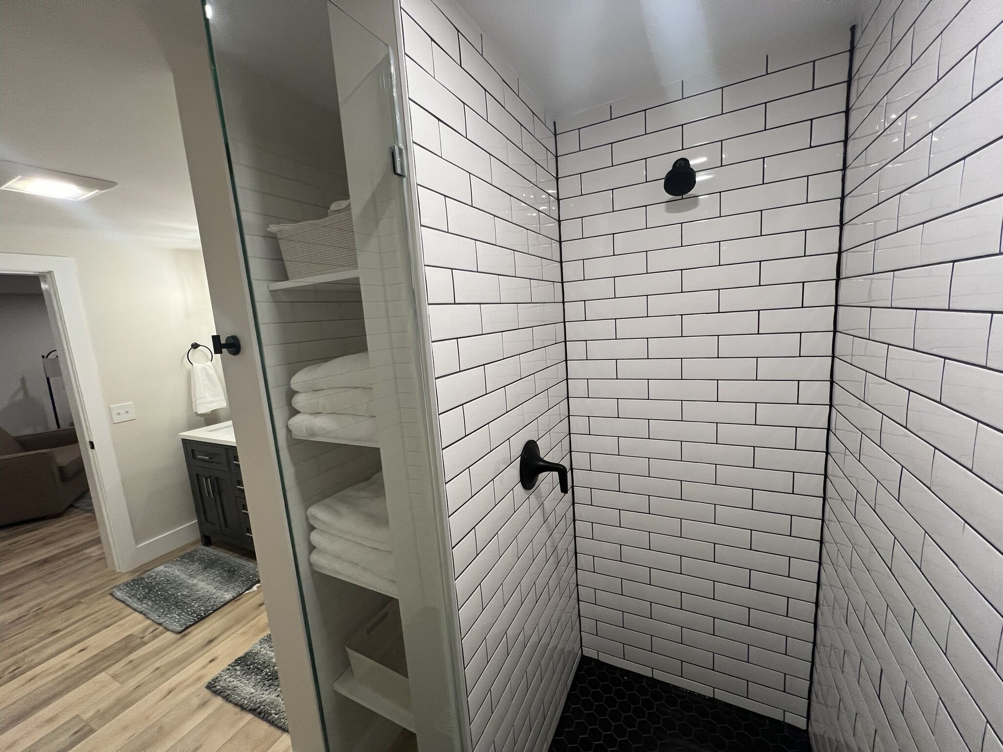 A bathroom with white subway tile walls, a glass shower enclosure, and a wooden floor.
