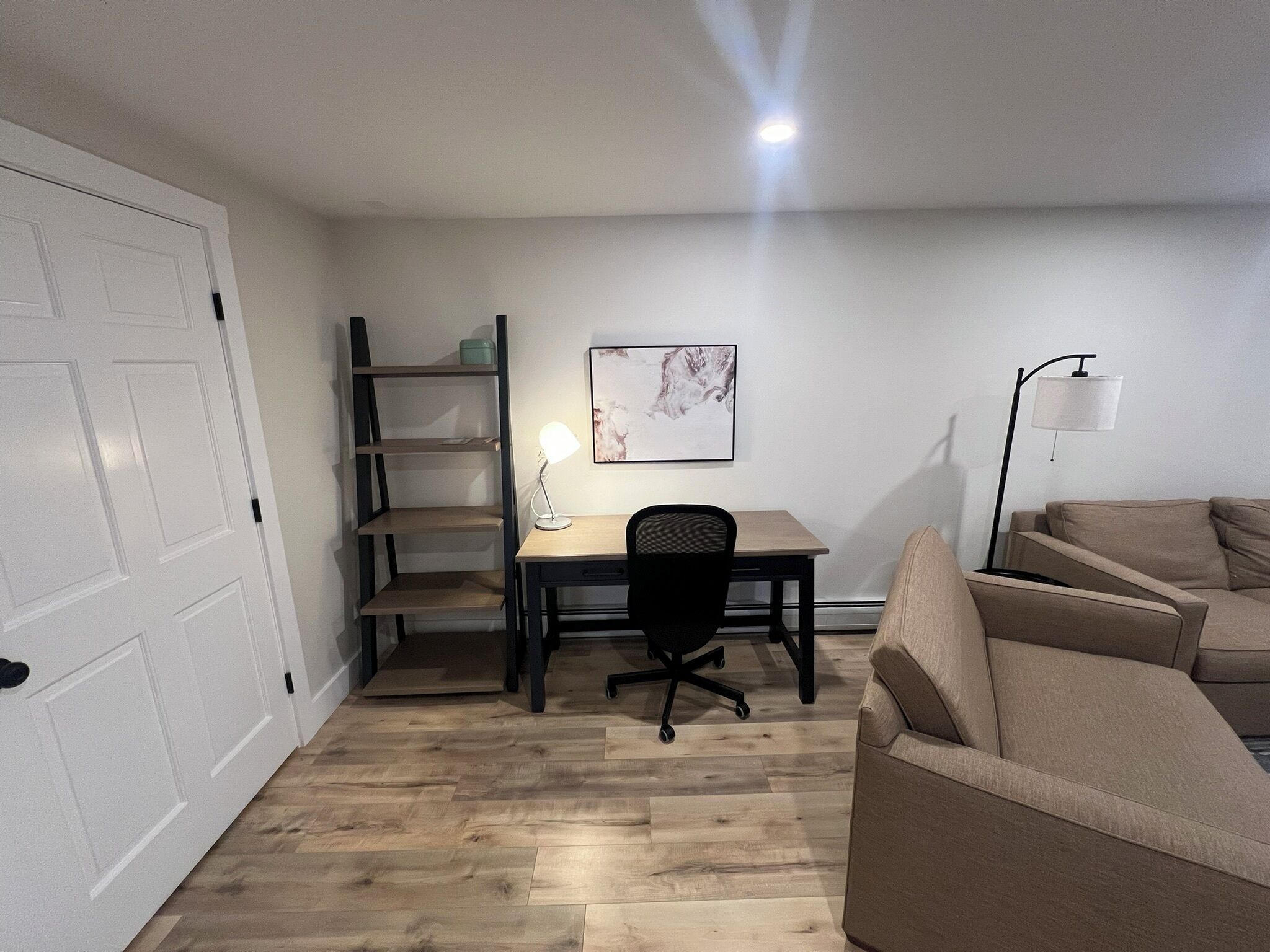 A room with a wooden floor, a desk with a chair, a bookshelf, a sofa, and a floor lamp.