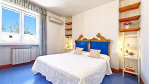 A bedroom with a large bed, a window with curtains, a radiator, and a shelf with decorative items. - La Bella Forianella. Alloro (Forio)