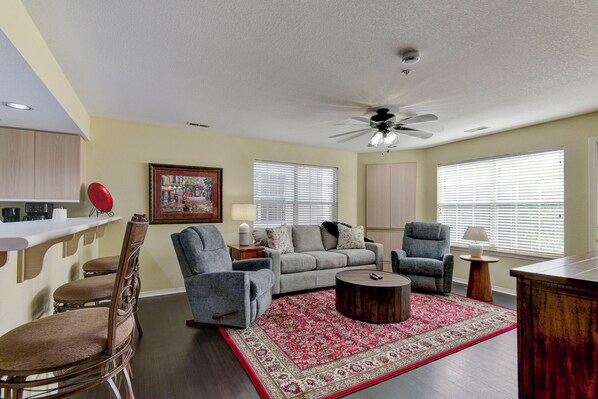 A living room with a grey sofa, two armchairs, a coffee table, and a red patterned rug. There is a ceiling fan, a framed picture on the wall, and a kitchen area with a counter and chairs.