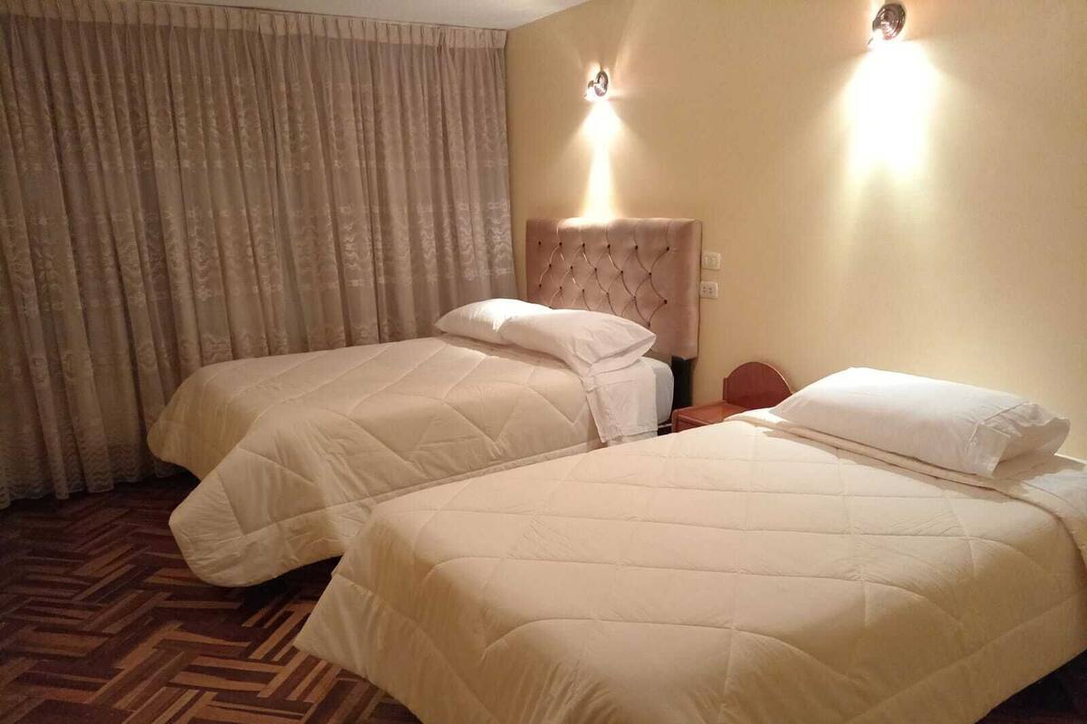 A hotel room with two beds, a wooden herringbone floor, and a large window with curtains.