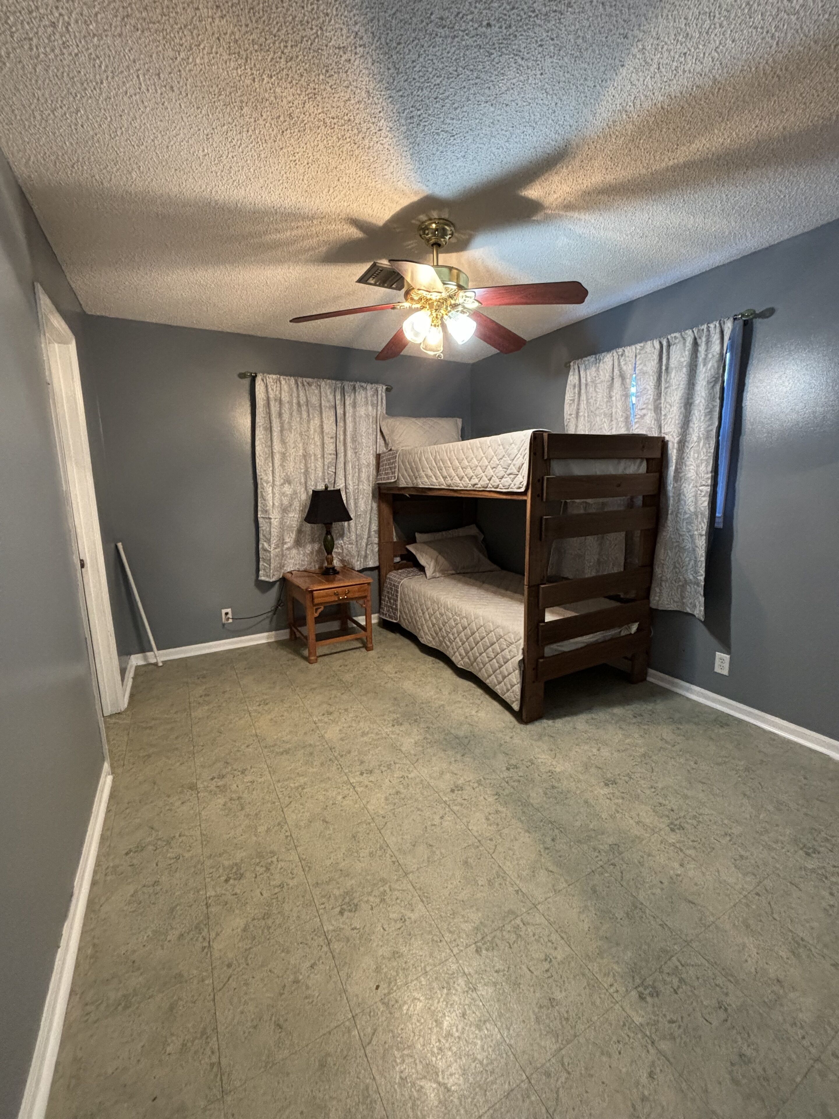 Une chambre avec un lit superposé, un ventilateur de plafond, une petite table de chevet en bois et une lampe.