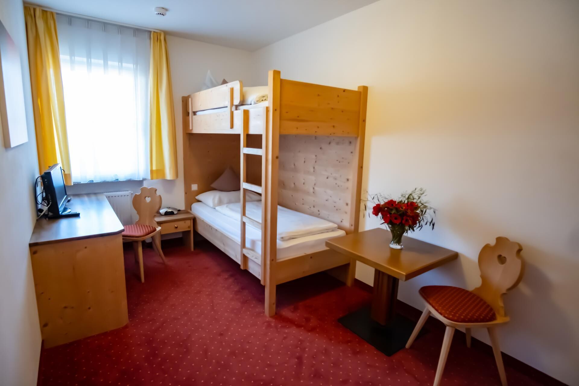 Ein Zimmer mit Etagenbett, Schreibtisch mit Laptop, Stuhl, kleinem Tisch mit Blumenvase und einem Fenster mit Vorhängen.