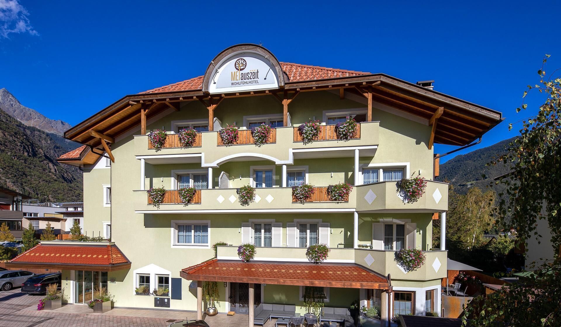 Un hôtel à plusieurs étages, de style architectural alpin traditionnel, doté de balcons ornés de jardinières, d’une enseigne indiquant qu’il s’agit d’un hôtel, et offrant une vue sur un paysage montagneux.
