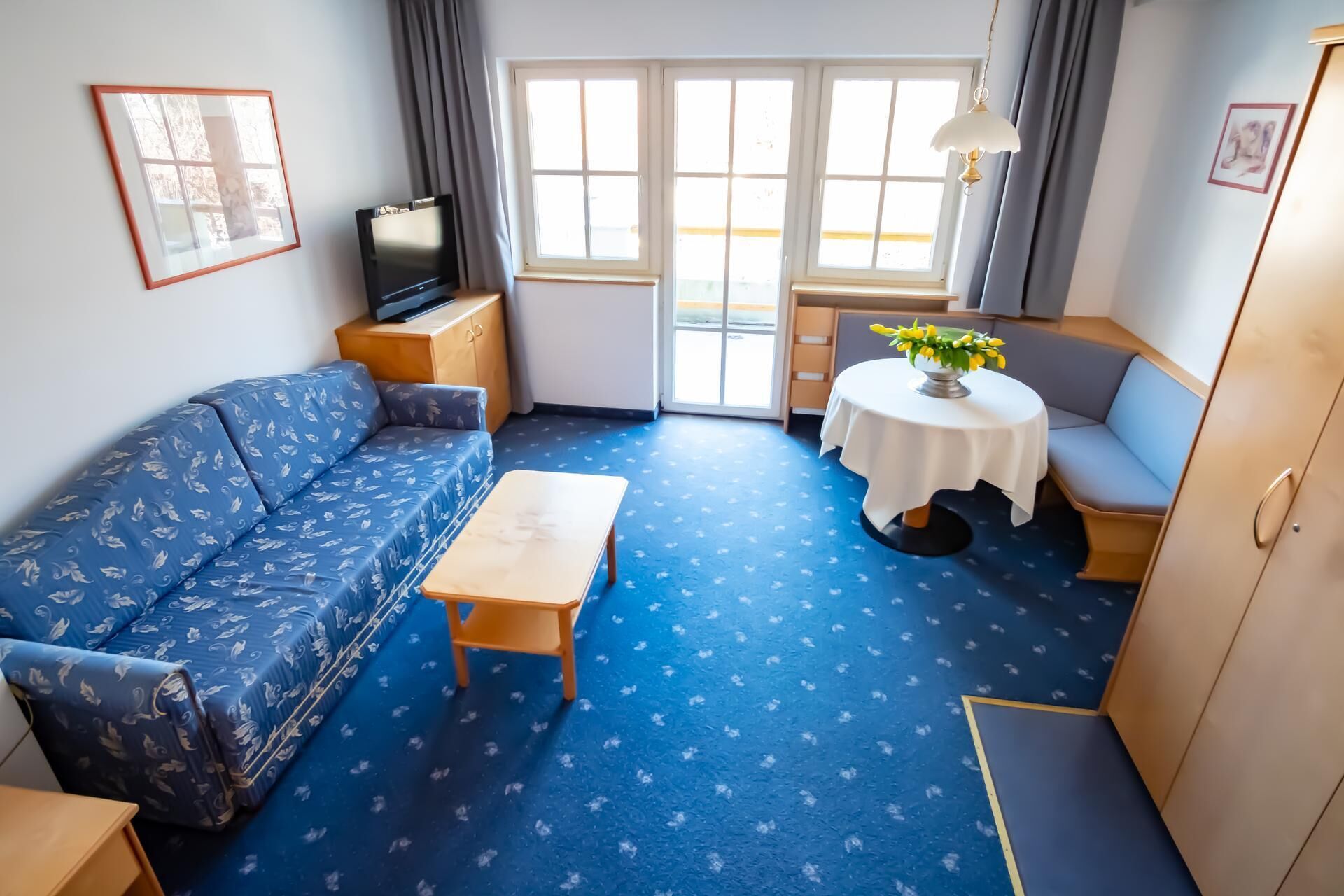 Ein gemütlicher Raum mit Sofa, kleinem Tisch, Fernseher, Sessel, einem kleinen Tisch mit Blumen und einem Kleiderschrank.