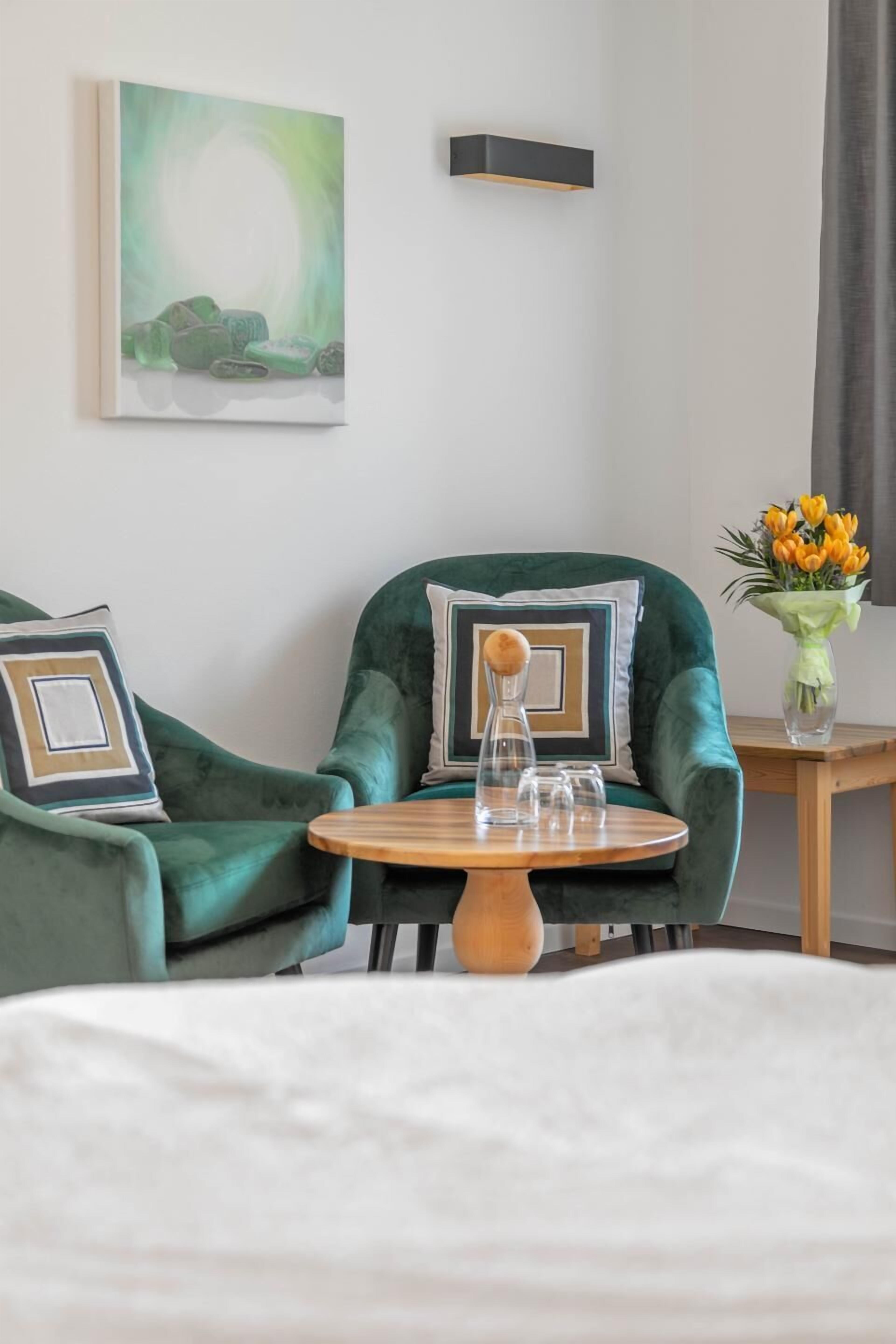 Habitación de hotel con un sillón verde, una mesita de madera, un cuadro en la pared y un jarrón con flores.