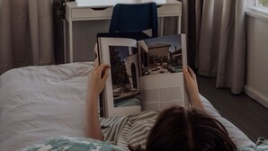 Iemand ligt op een bed te lezen in een kamer met een groot raam, gordijnen en een hangplant.
