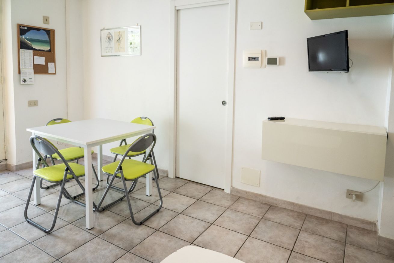 A room with a white table and four green chairs, a white door, a bulletin board with a picture and notes, a mounted television, and a shelf with a remote control.