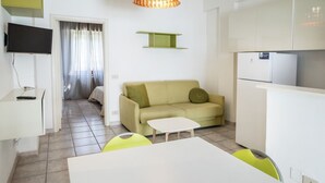 A modern, well-lit room with a sofa, a small table, a TV, and a kitchen area with a refrigerator. - M377 - Marcelli, two-room apartment with external courtyard and parking (Marcelli)