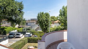 Un balcón con una silla blanca, una mesita y vistas a una zona residencial con casas y coches aparcados.