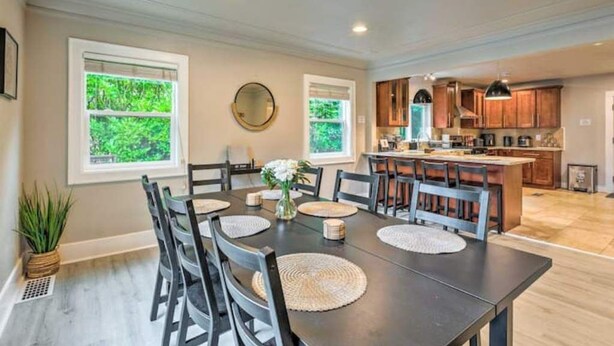 A dining area with a large table, chairs, and a vase of flowers. There are windows with greenery visible outside.