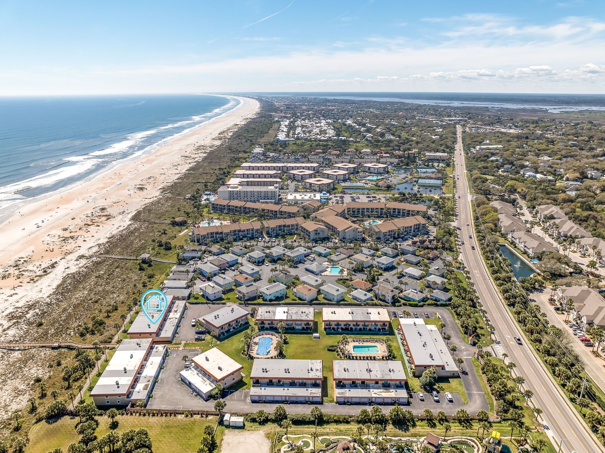 An aerial view of a coastal resort with multiple buildings, a beach, and a road.