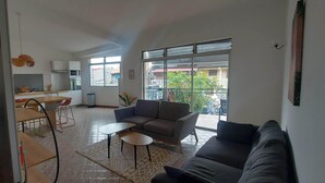 A modern living room with a sofa, a coffee table, and a dining area with a table and chairs. There is a kitchen in the background with cabinets and appliances. - Appartement Spacieux et Lumineux à 20m de la Plage ! (Les Anses-d'Arlet)