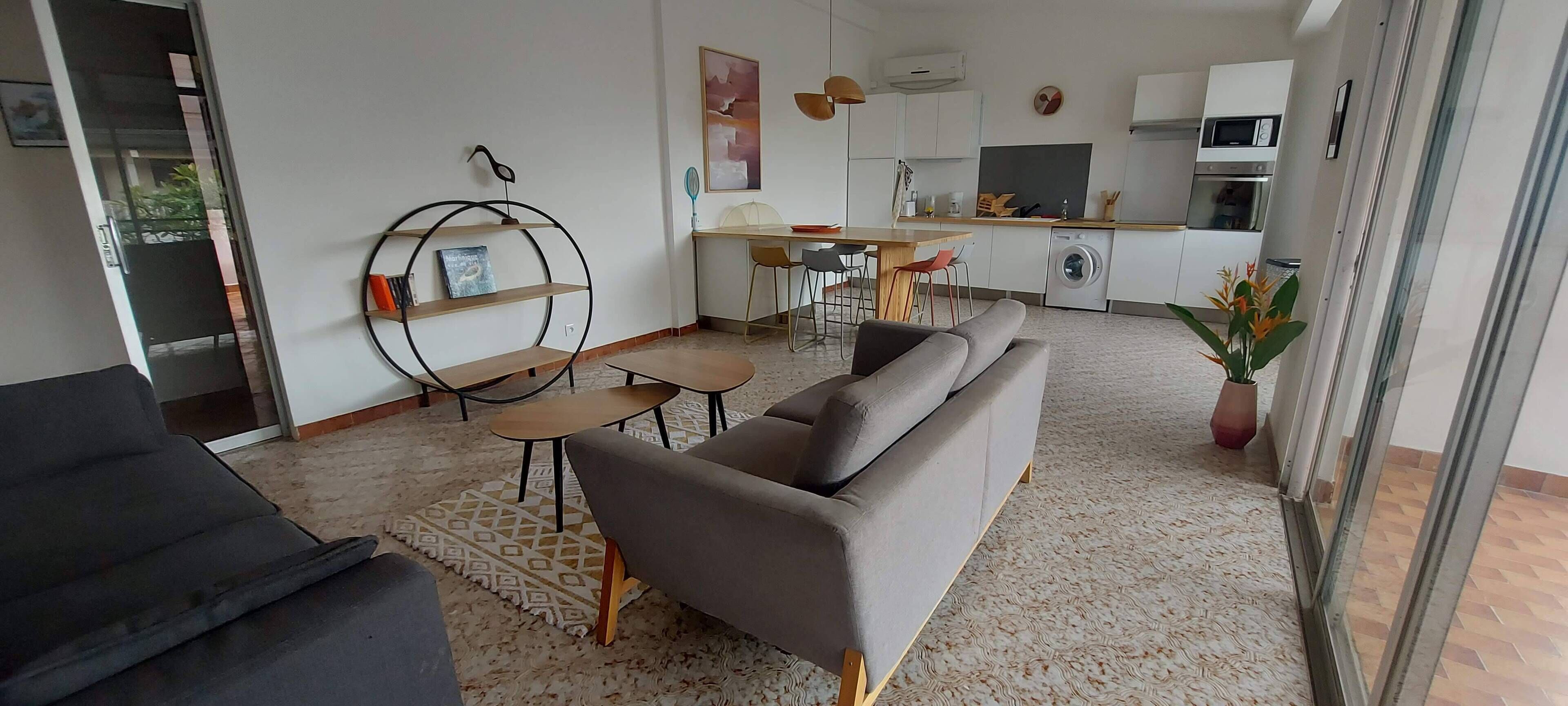 A modern living room with a grey sofa, a small round table, and a coffee table. There is a kitchen area with a dining table and chairs, a washing machine, and a microwave.