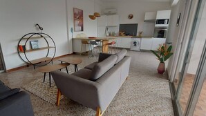 A modern living room with a grey sofa, a small round table, and a coffee table. There is a kitchen area with a dining table and chairs, a washing machine, and a microwave. - Appartement Spacieux et Lumineux à 20m de la Plage ! (Les Anses-d'Arlet)