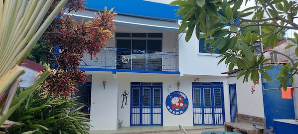 A two-story building with blue doors and windows, a balcony with decorative railings, and a circular sign with a cartoon character. - Appartement Spacieux et Lumineux à 20m de la Plage ! (Les Anses-d'Arlet)
