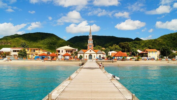 A pier leading to a coastal town with a church, beach houses, and mountains in the background. - Appartement Spacieux et Lumineux à 20m de la Plage ! (Les Anses-d'Arlet)