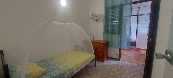A bedroom with a single bed, a wooden dresser, a wall fan, and a window with blinds. - Appartement Spacieux et Lumineux à 20m de la Plage ! (Les Anses-d'Arlet)