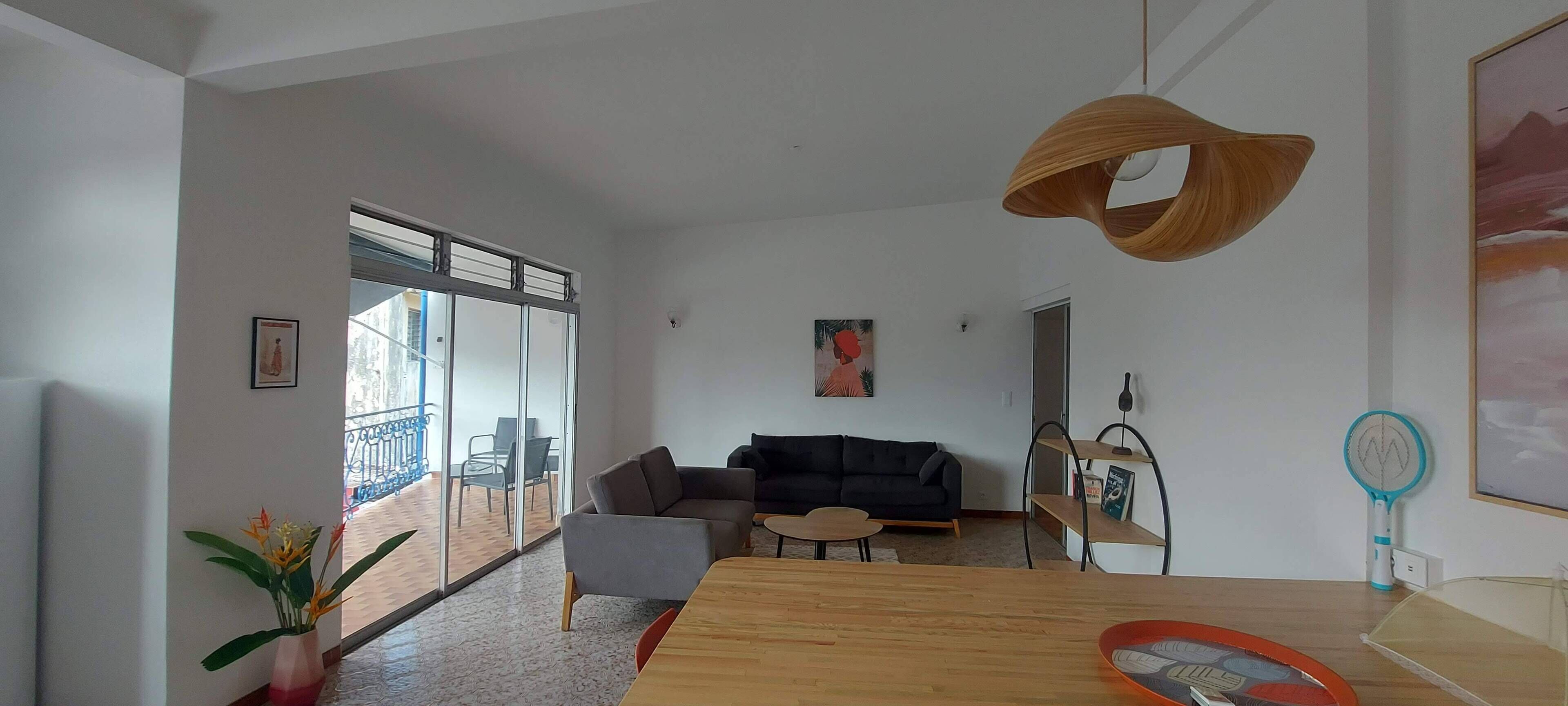 A modern living room with a wooden dining table, a black sofa, and a glass door leading to a balcony.