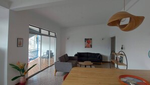 A modern living room with a wooden dining table, a black sofa, and a glass door leading to a balcony. - Appartement Spacieux et Lumineux à 20m de la Plage ! (Les Anses-d'Arlet)