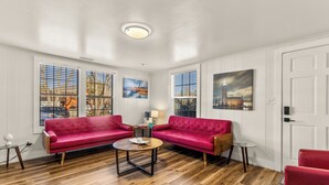 Un salon spacieux avec deux canapés rouges, une table basse ronde et une petite table d’appoint. De grandes fenêtres équipées de stores laissent entrer la lumière naturelle et offrent une vue sur le paysage urbain.