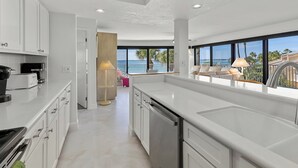 Una cocina moderna con muebles blancos, lavavajillas y una isla grande. Se ve el océano a través de ventanas amplias.