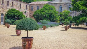 Un patio con suelo de grava, rodeado de edificios de piedra y árboles topiarios en macetas grandes.