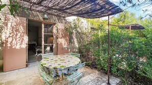 Un patio exterior con una mesa redonda y sillas, una pérgola que proporciona sombra, y vistas de un jardín con diversas plantas.