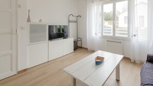 Ein moderner, hell erleuchteter Raum mit einem Fernseher auf einem weißen Schrank, einem kleinen Tisch mit einem blauen Gegenstand und einem Fenster mit durchsichtigen Vorhängen.