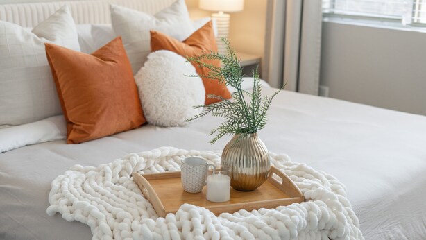 Een netjes opgemaakt bed met een witte deken, een witte gebreide plaid, en een dienblad met een vaas met groen, een mok en een glas melk.
