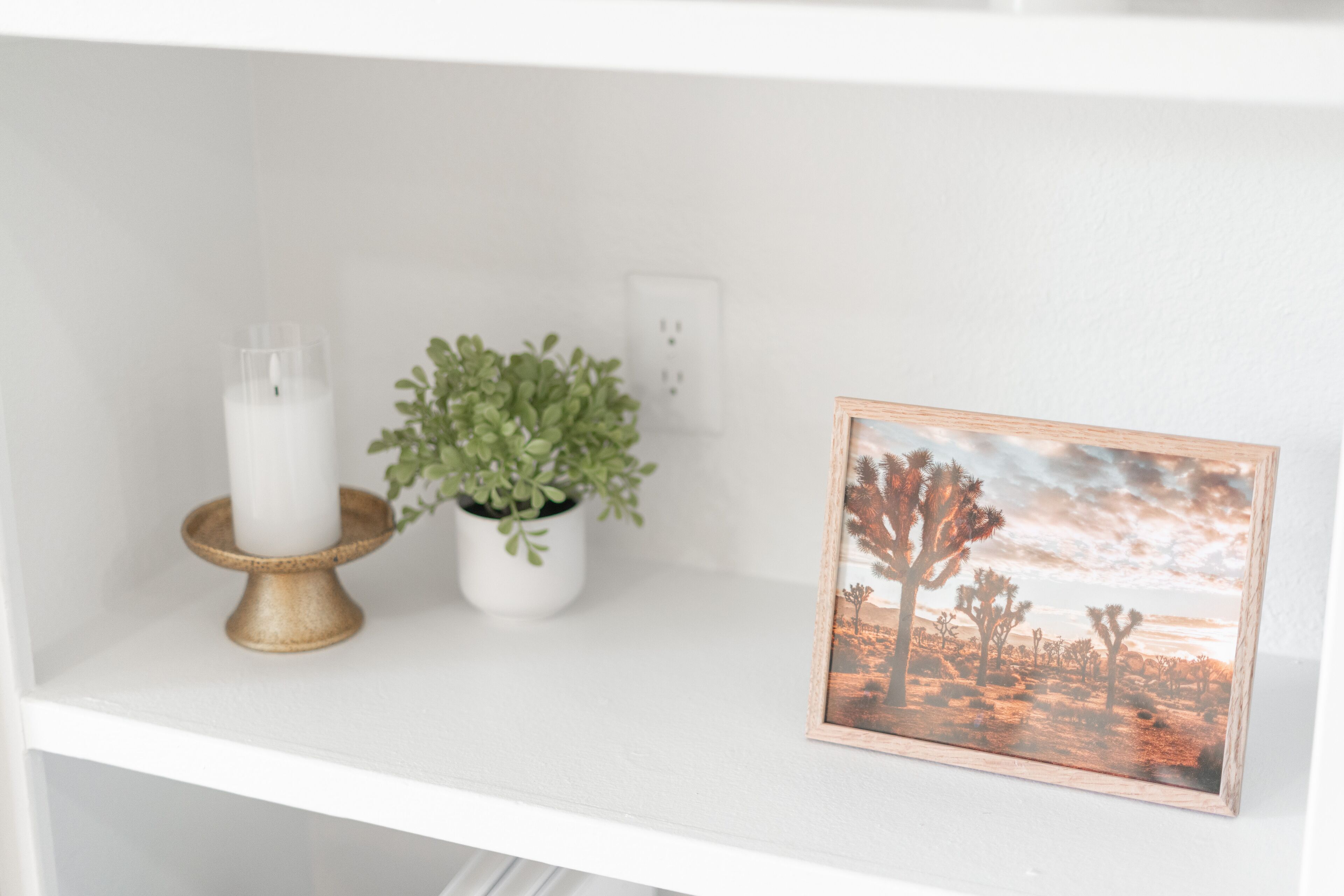 A white shelf with a white candle, a small potted plant, and a framed picture of a desert landscape.
