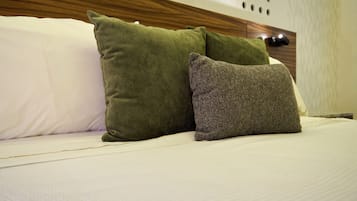 Un lit avec une literie blanche et deux oreillers, un vert et un gris, dans une pièce dotée d’une tête de lit en bois et d’un mur blanc orné d’ouvertures circulaires.