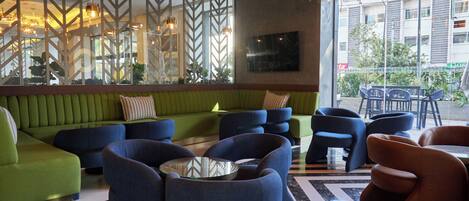 A modern lounge area with green and blue seating, a glass coffee table, and a view of the cityscape through large windows.