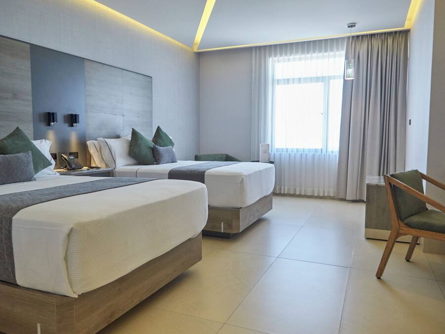 Quarto de hotel moderno com duas camas, uma cadeira de madeira e uma janela grande com cortinas.