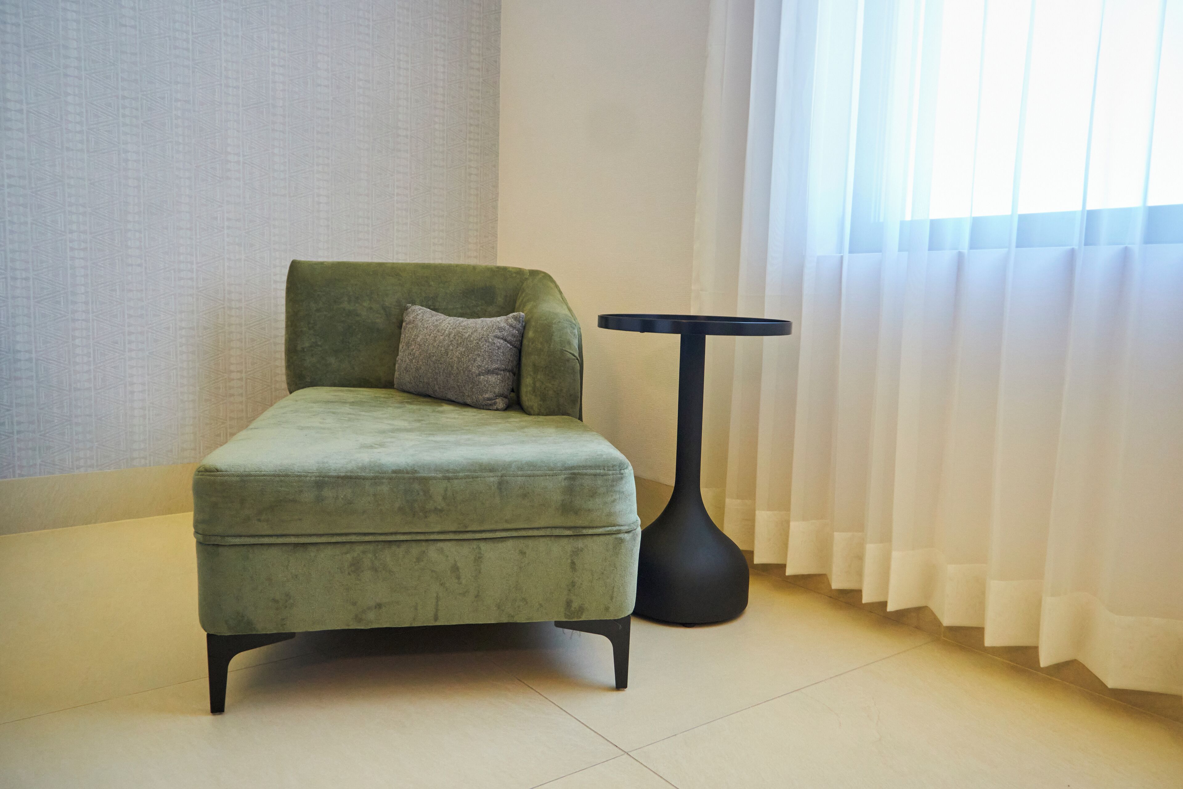 Un sillón verde con un cojín, una lámpara de pie negra y cortinas blancas en una habitación con pared estampada y suelo de baldosas.