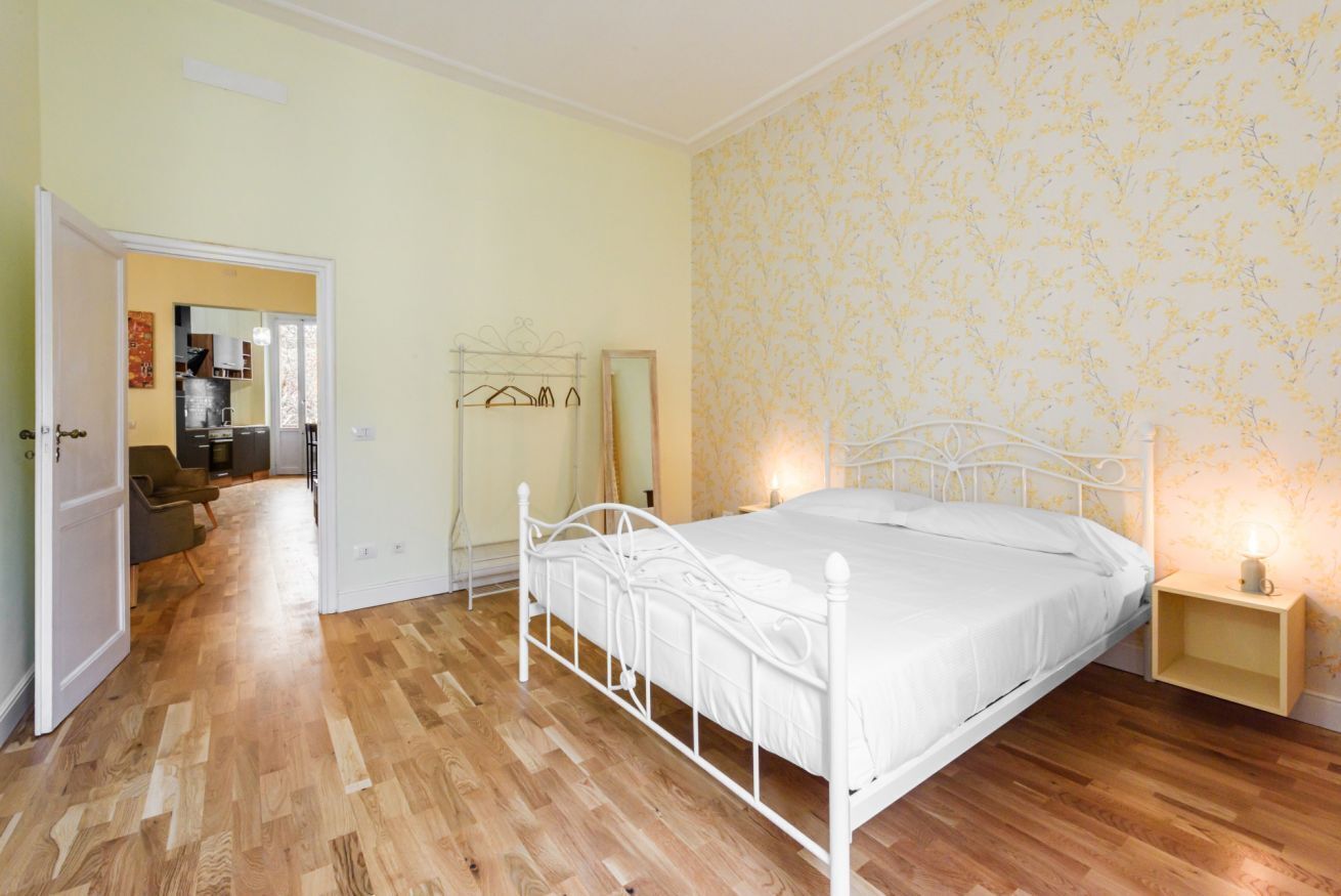 Un dormitorio con estructura de cama de metal blanco, suelo de madera, papel tapiz floral y una mesita de noche con lámpara.