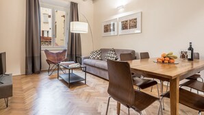 A modern living room with a wooden dining table, leather chairs, a sofa with patterned cushions, a floor lamp, and artwork on the wall.