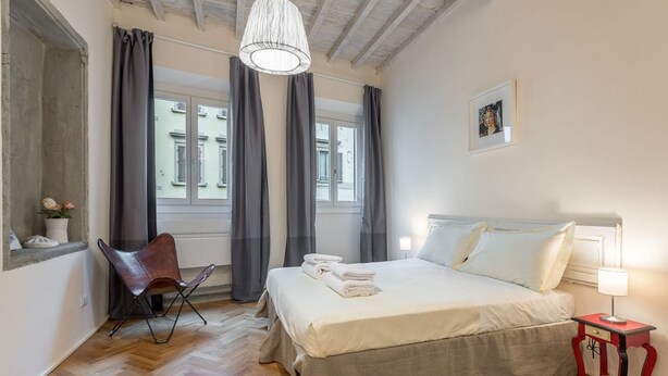 A bedroom with a bed, a chair, a nightstand, a lamp, a window, and curtains.